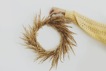 Autumn boho wreath. Hand in yellow sweater holding stylish rustic wreath with dry grass on white wall background. Making rustic autumn wreath with pampas grass, holiday workshop