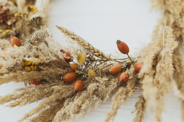 Stylish autumn rustic wreath close up. Creative boho wreath with dried pampas grass, tansy wildflowers, wheat, dog-rose berries on white wooden table. Fall decor details