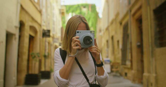 Young happy woman make photo on instant camera that print out photo in real time, look at picture and smile. Authentic and candid excited woman travel on road trip in european village