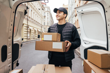 Male courier in uniform standing at van trunk holding boxes preparing for delivery to customer - obrazy, fototapety, plakaty