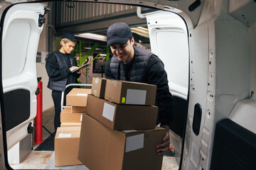 Smiling male courier loads cardboard boxes from a cart into the car while coworker checking...