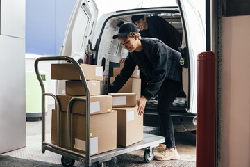 Female courier loads cardboard boxes from a cart into a van while a colleague stands in it