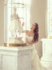Beautiful woman with long hair in a light dress and a big doll ballerina in the theater under a glass dome