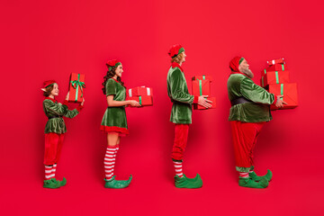Full body photo of line santa service people different age hold giftboxes wear new year costumes isolated on red background