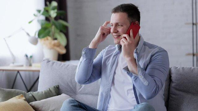 Portrait of joyful male resting at home sitting on sofa in modern room speaking on smartphone. Young man talking on mobile phone. Caucasian cheerful Guy using phone indoor. Conversation concept.