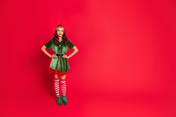 Full size photo of cool brunette santa helper young lady stand near empty space wear costume isolated on red color background