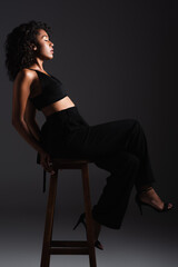 full length of young african american woman in sexy crop top and pants sitting on stool on black