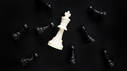 The white chess king lies on the table surrounded by black pawns. Chess pieces on a black background.