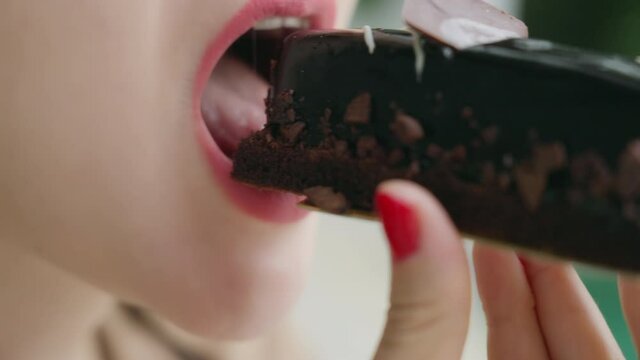 Close-up of a girl with red lipstick lips biting eclair covered with dark chocolate. A woman with a red manicure has a delicious chocolate dessert against a blurry white-green background. Slow motion.