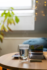 Glass cup of hot water on wooden table with watercolor drawing in paper notebook near home flower and bed in bedroom near window and lights on cozy cloudy autumn or winter day. Drink, weekend, holiday