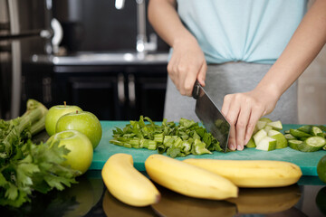 Close-up of unrecognizable woman standing at counter with vegetables and fruits and cutting celery on plastic board
