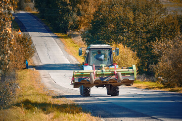 Fototapeta premium Tractor with plough rides along rural asphalt road. Tractor and rolling hills, agricultural machinery, farming concept. Tractor with plow driving on hilly road at countryside, agricultural autumn work