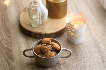 Bowl of cinnamon cookies, cup of warm drink, lit candles and vase with gypsophila flowers on the table. Selective focus, fairy lights in the background. Hygge at home.