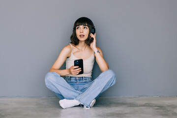 Portrait of a cheerful woman looking away while sitting on a floor with headphones and holding mobile phone over gray background