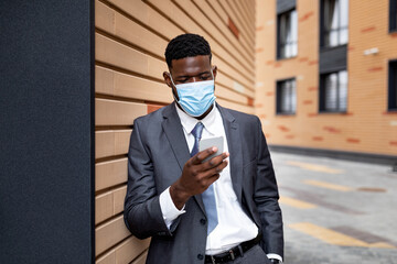 African american entrepreneur in protective face mask using smartphone, standing outdoors near office building