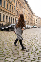 Portrait of young brunette caucasian woman dressed casually in brown hoodie and coat walking on street near residential houses and cars on cloudy early autumn day. Holiday, leisure, lifestyle