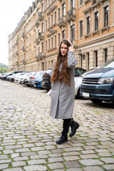 Fototapeta premium Portrait of young brunette caucasian woman dressed casually in brown hoodie and coat walking on street near residential houses and cars on cloudy early autumn day. Holiday, leisure, lifestyle