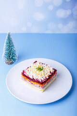 Herring salad under a fur coat. Traditional Russian multilayered salad from herring, beets, potatoes, carrots and eggs. Close-up, blue background, miniature Christmas tree.