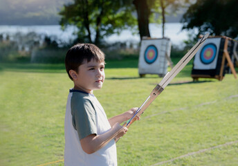 Portrait Young kid practising his archery, Mixed race child doing outdoors activity in summer camp adventure. School boy shooting bow with archery on training in sport school. - obrazy, fototapety, plakaty