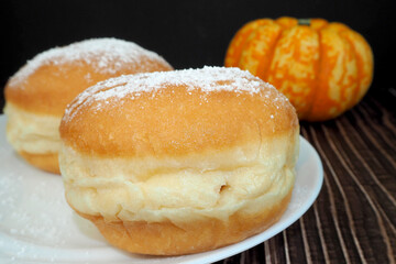 two round donuts sprinkled with powdered sugar stand on a white plate against a background of a small pumpkin on a dark background