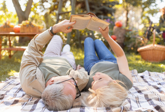 Married senior couple reading book together while lying on picnic blanket in garden, spending free time outdoors