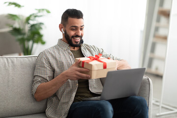 Arab guy celebrating holiday online, holding gift box during video call with friend, sitting on sofa with laptop at home