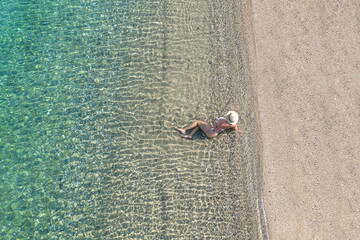 Young woman in hat, swimsuit sitting in the water on the shore of sea on the sand beach. Summer. View from above. Top view, copter