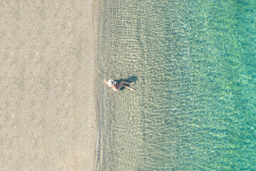 Young woman in hat, swimsuit sitting in the water on the shore of sea on the sand beach. Summer. View from above. Top view, copter