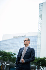 Confident mature Caucasian businessman in formal suit and glasses standing against skyscraper outdoors