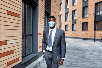 Confident african american businessman walking outdoors near business center, wearing protective face mask