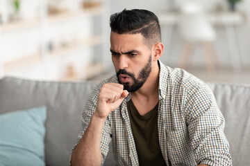 Coronavirus, influenza concept. Sick young Arab man coughing into fist, sitting on couch, feeling...