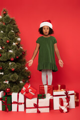 Amazed african american kid in santa hat pointing at presents near christmas tree on red background