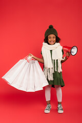 Cheerful african american girl in scarf holding shopping bags and loudspeaker on red background