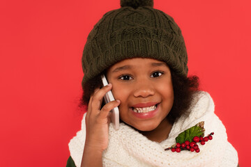 Happy african american girl in hat and scarf with mistletoe talking on mobile phone isolated on red