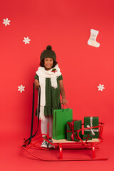 Smiling african american child in scarf with mistletoe standing near sleigh, presents and shopping bag on red background