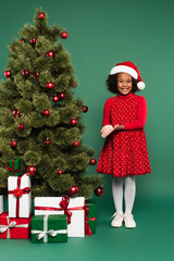 Smiling african american girl in santa hat pointing at presents under christmas tree on green background