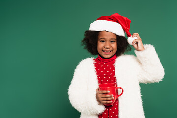Smiling african american child in santa hat and fluffy jacket holding red cup isolated on green