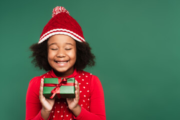 Joyful african american kid in warm hat closing eyes while holding gift box isolated on green
