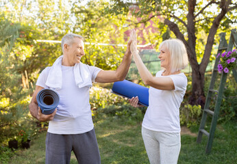 Obraz premium Active mature couple standing with yoga mats and giving high five each other after training in garden outdoors