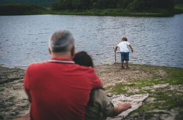Fototapeta premium a little boy is playing by the water and his parents are watching him in the foreground on a cloudy day, rear view the average plan