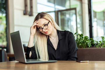 Tired blonde business woman looks into the laptop screen while sitting at a table in an outdoor cafe. Freelance, remote work