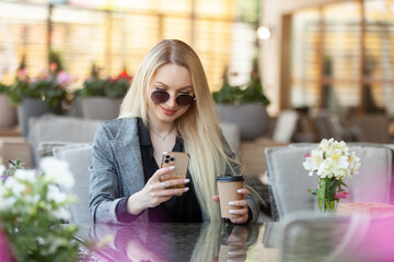 Young blonde woman in sunglasses holds cup of coffee and uses smartphone while sitting at table in summer terrace of cafe