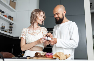 Handsome man makes a surprise to his lovely woman while she is cooking in the kitchen. The husband gives a box with a gift to his wife. Romantic love concept. Birthday or anniversary
