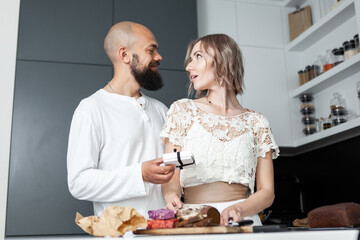Handsome man makes a surprise to his lovely woman while she is cooking in the kitchen. The husband gives a box with a gift to his wife. Romantic love concept. Birthday or anniversary