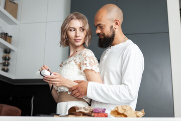 Handsome man makes a surprise to his lovely woman while she is cooking in the kitchen. The husband gives a box with a gift to his wife. Romantic love concept. Birthday or anniversary