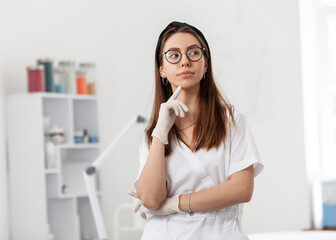 Pensive professional doctor cosmetologist dermatologist woman in her beauty salon in white medical clothes