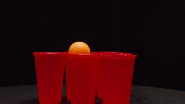 Person throws small ball into red plastic cups with drinks playing beerpong on table in dark studio extreme close view