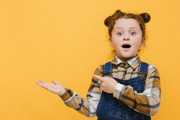 Portrait of shocked little caucasian child girl points fingers aside, wears shirt, posing isolated over yellow color background wall in studio with copy space. Childhood emotion lifestyle concept