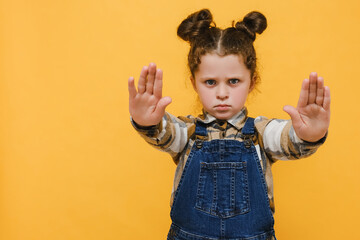 Portrait displeased little kid girl demonstrate stop gesture, wears shirt, posing isolated over yellow color background studio with copy space for advertisement. Childhood emotional lifestyle concept