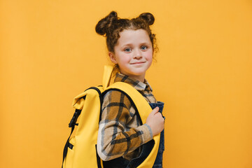 Portrait of cute beautiful happy smiling caucasian little girl, child schoolgirl holding backpack standing over yellow studio background wall, positive looking at camera. Back to school concept